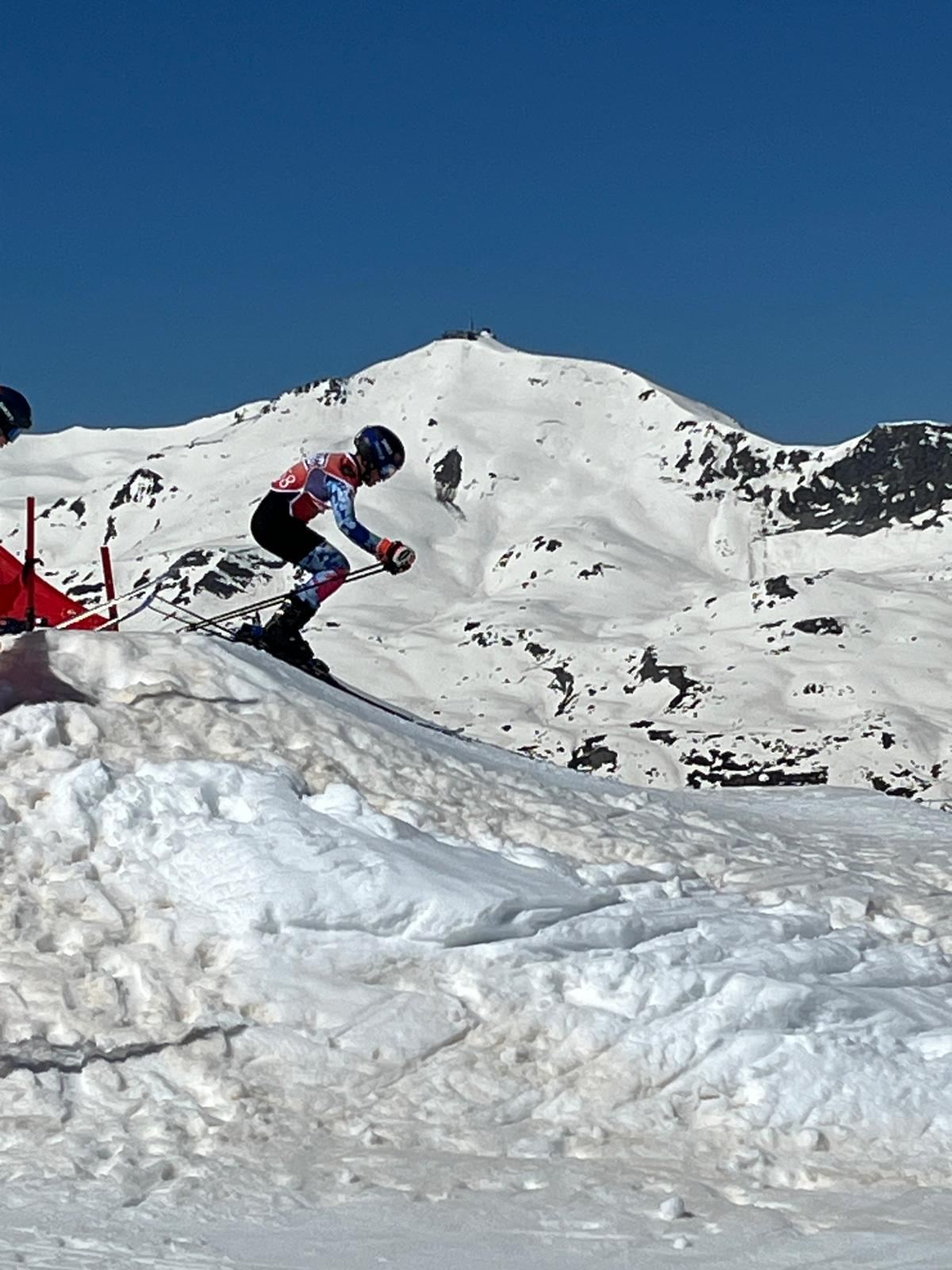 Deportista en el aire sobre un salto de skicross, con una montaña nevada y el cielo azul de fondo.
