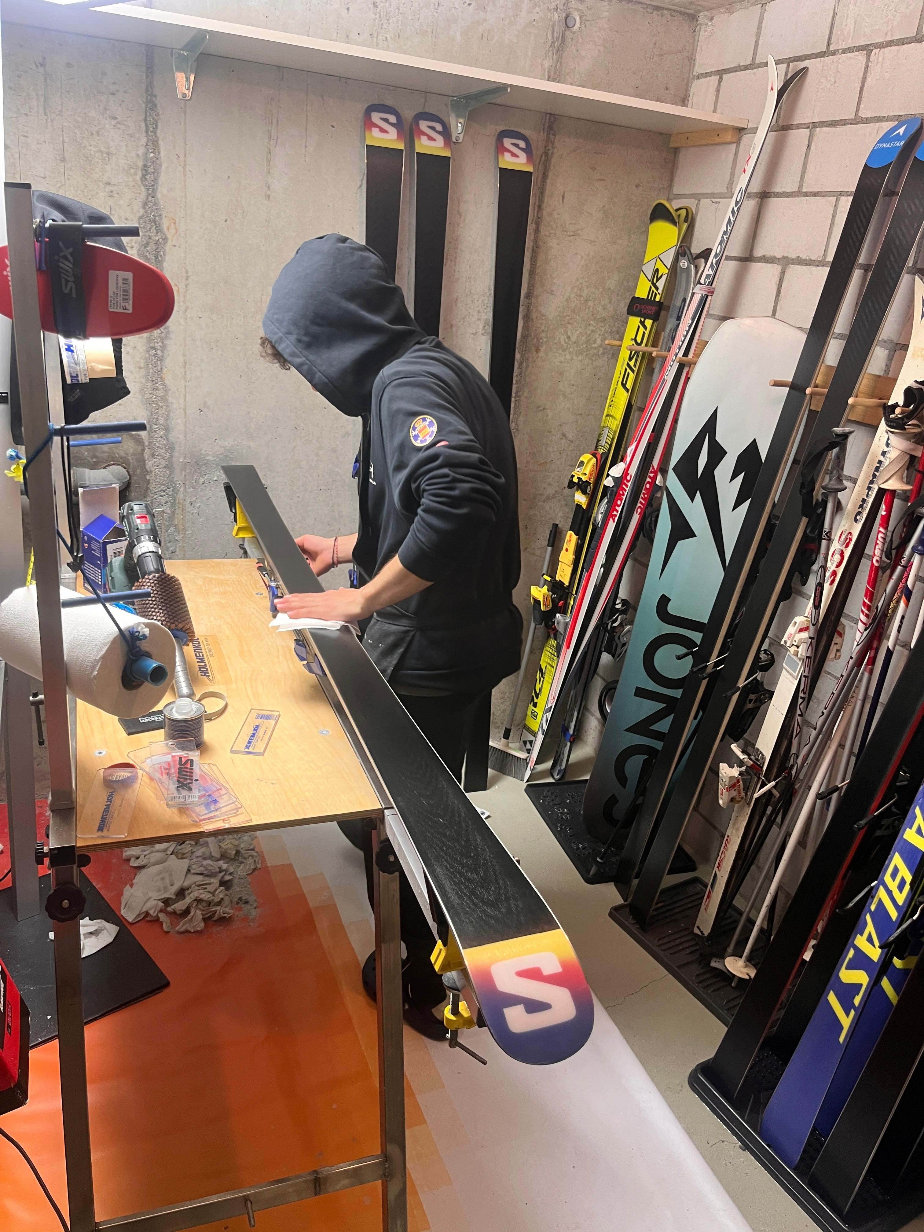 Un skiman prepara unos esquís en el taller, rodeado de esquís y herramientas.