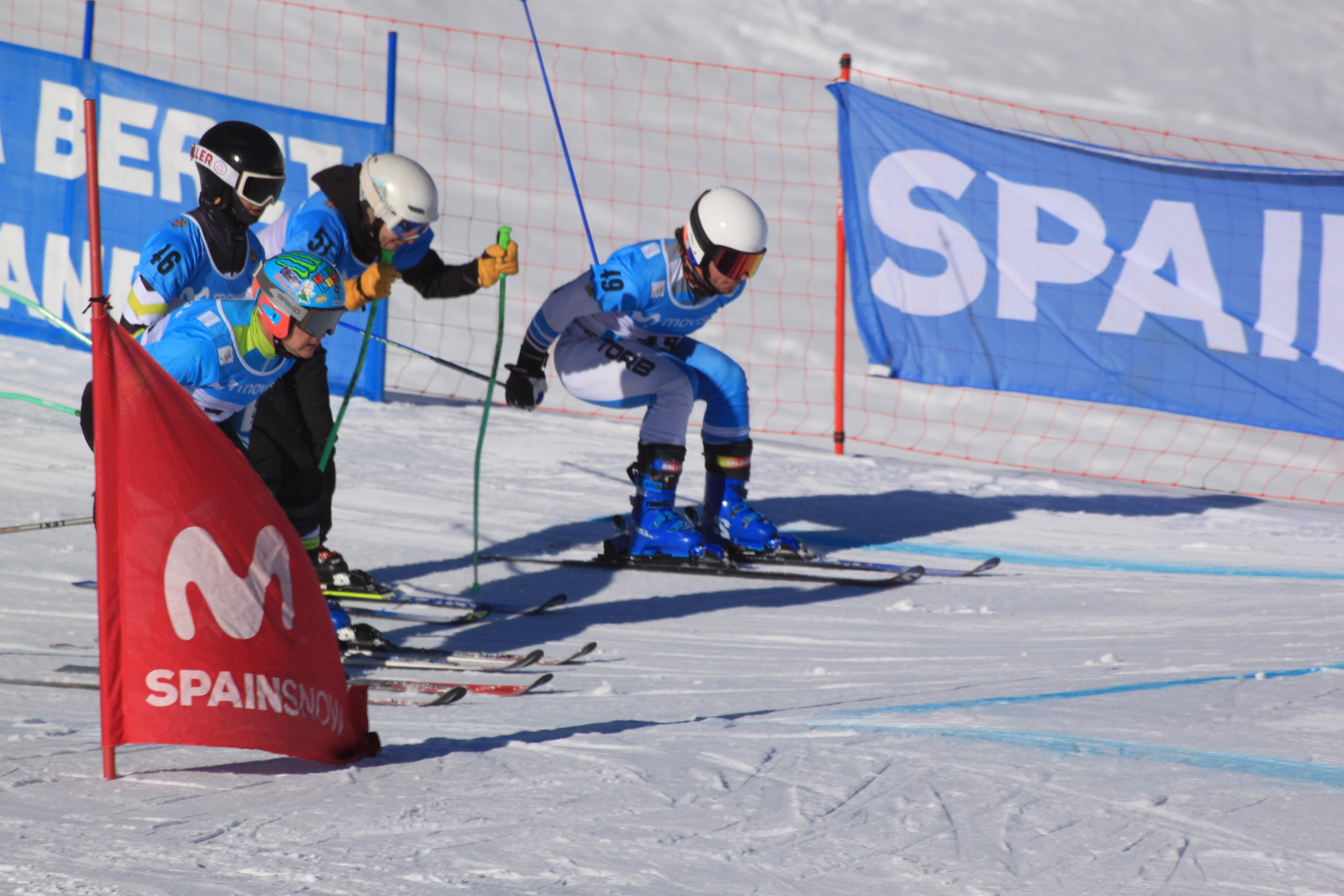 Tres corredores con dorsal salen juntos en una manga de skicross, junto a la bandera de la RFEDI.