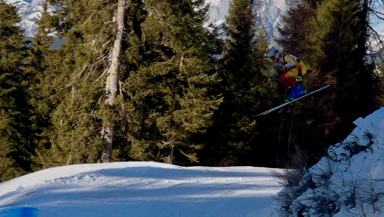Un corredor salta en un circuito de skicross entre abetos, con montañas nevadas al fondo.
