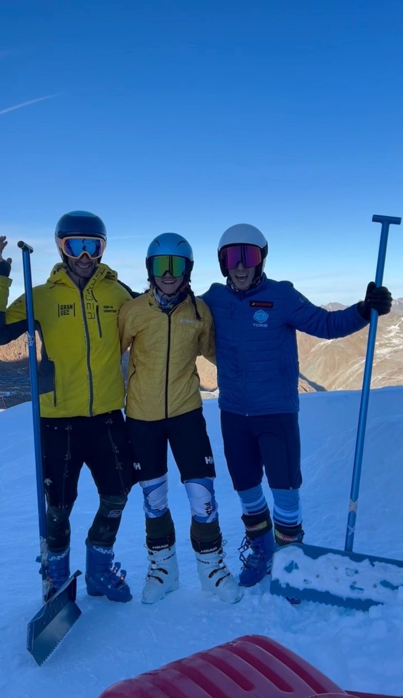 Tres personas del equipo sonríen con palas en la mano sobre el circuito de Pitztal, con el cielo azul y las montañas detrás.