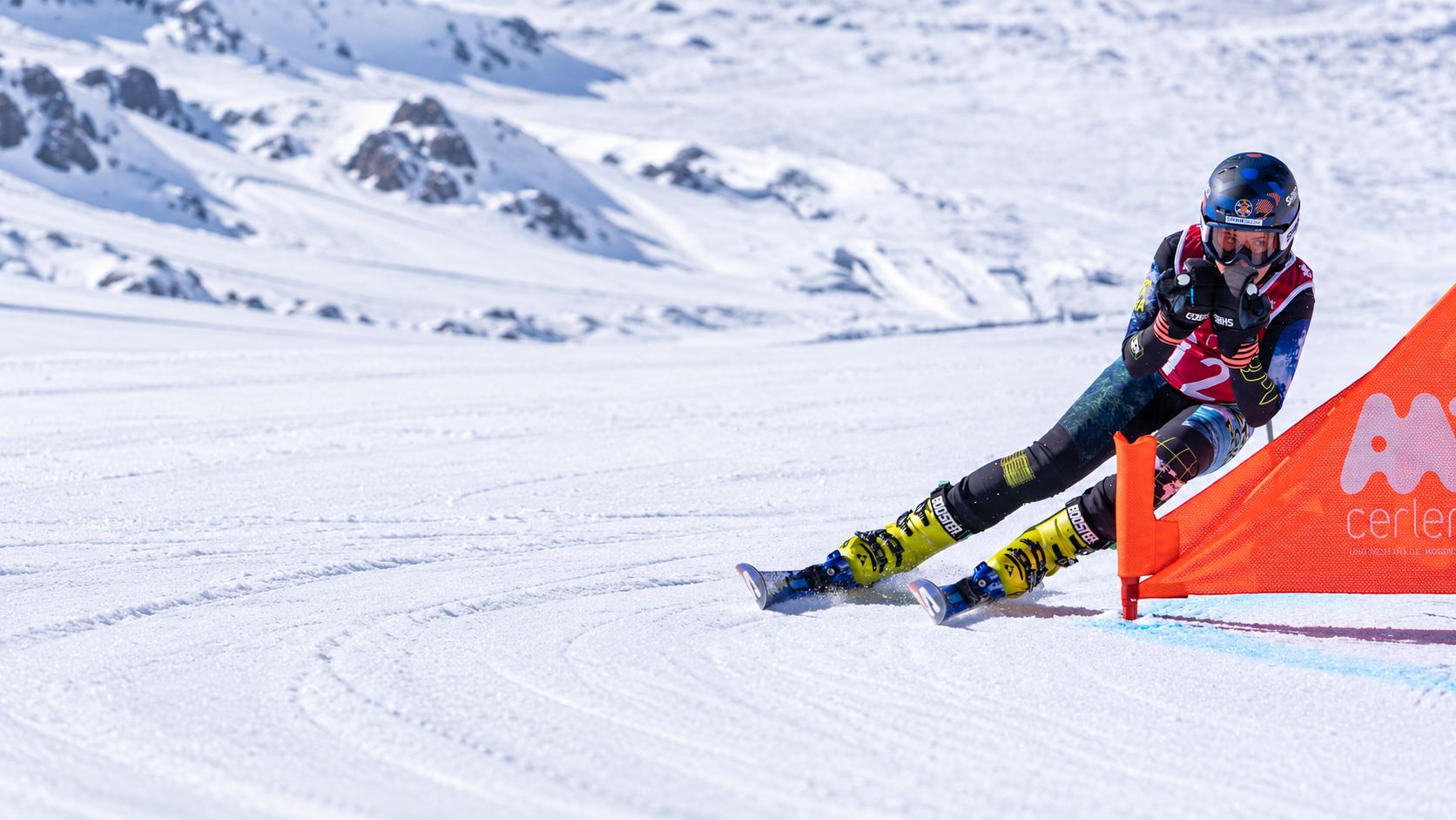 Deportista del equipo trazando una curva junto a una puerta naranja de Cerler, con montañas nevadas al fondo.